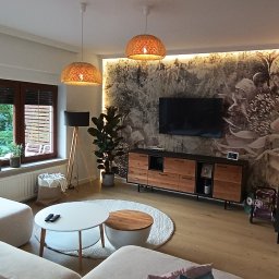 StyleHouse - Jasny salon z drewnianą podłogą, sofą w odcieniach bieli, okrągłym stolikiem kawowym i ścianą z motywem kwiatowym podświetloną LED, dwie lampy wiszące z abażurami z plecionej wikliny.
