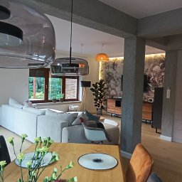 StyleHouse - Przestronne wnętrze domu z jasnym parkietem, szarą sofą, przeszklonymi lampami wiszącymi i kwiatami na drewnianym stole jadalnym, widok na salon i dekoracyjną tapetę w tle.