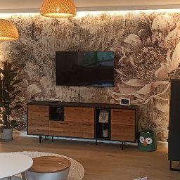 StyleHouse - Nowoczesny salon z tapetą w motywy kwiatowe, drewnianą komodą pod telewizorem, lampami z abażurami z wikliny i zielonym koszem w kształcie sowy.