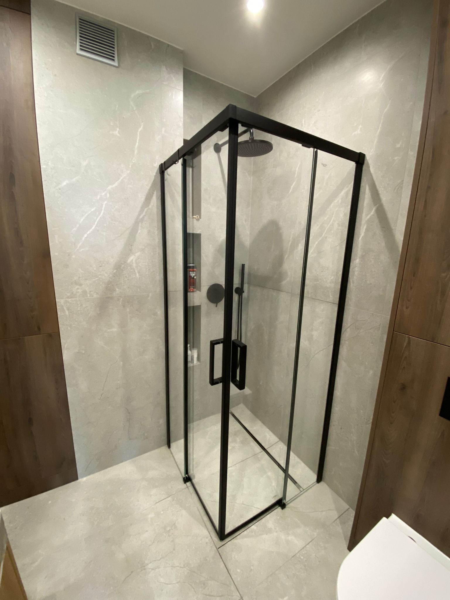 Nowoczesna kabina prysznicowa z czarnymi profilami i deszczownicą w łazience wykończonej płytkami imitującymi marmur, widoczny fragment toalety.