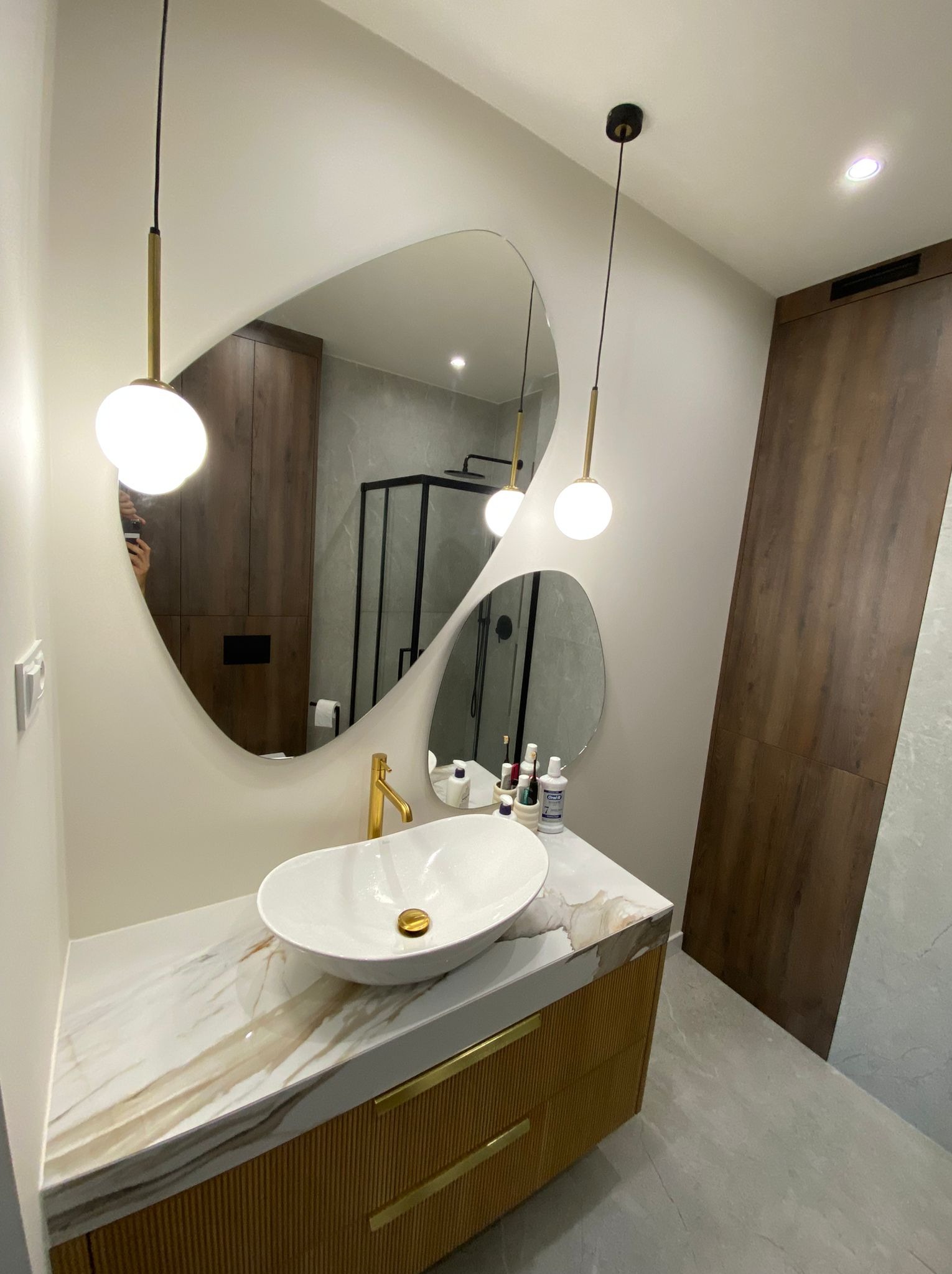 Nowoczesna łazienka z umywalką nablatową w kształcie łzy, złotą baterią i nieregularnym lustrem podświetlonym wiszącymi lampami kulami, w tle kabina prysznicowa z czarnymi profilami.