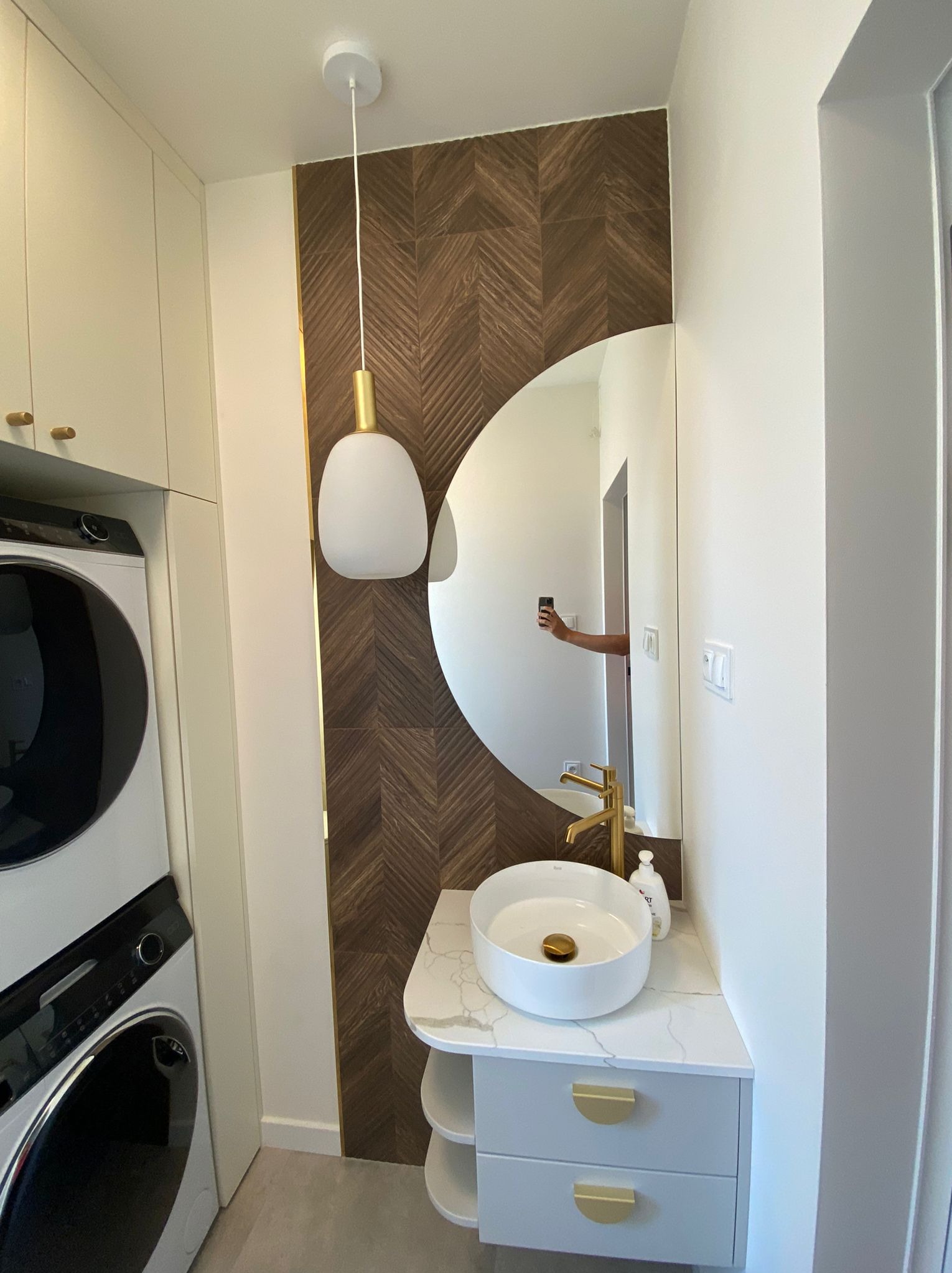 Nowoczesna łazienka z umywalką nablatową, złotą armaturą i dekoracyjną ścianą z płytek ułożonych w jodełkę, oświetlona wiszącą lampą z białym kloszem, obok pralka i suszarka w zabudowie.