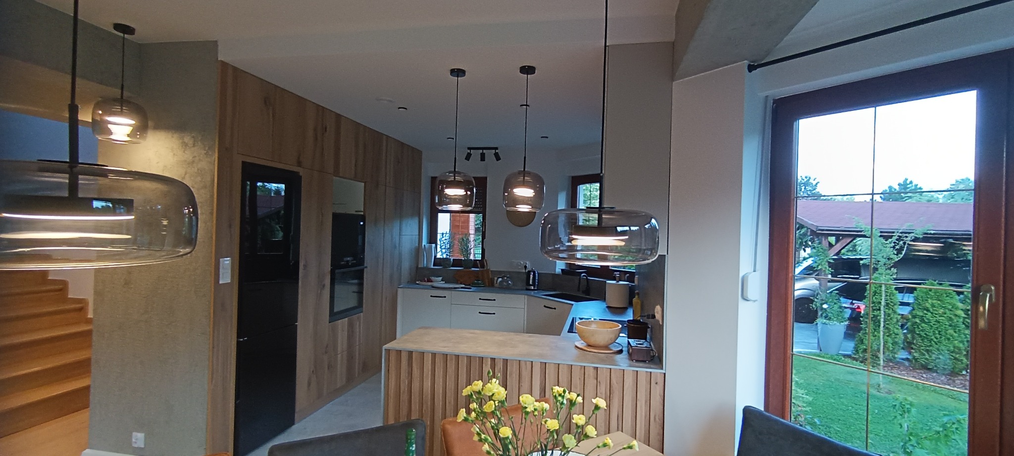 Nowoczesna kuchnia z wyspą, oświetlona wiszącymi lampami o dymionym szkle, widok na schody i ogród przez okno, drewniane wykończenie szafek.