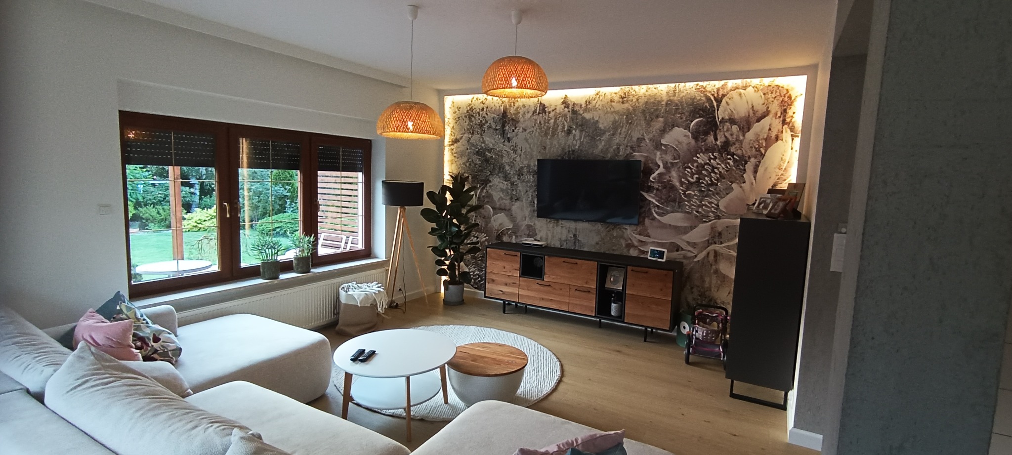 Jasny salon z drewnianą podłogą, sofą w odcieniach bieli, okrągłym stolikiem kawowym i ścianą z motywem kwiatowym podświetloną LED, dwie lampy wiszące z abażurami z plecionej wikliny.