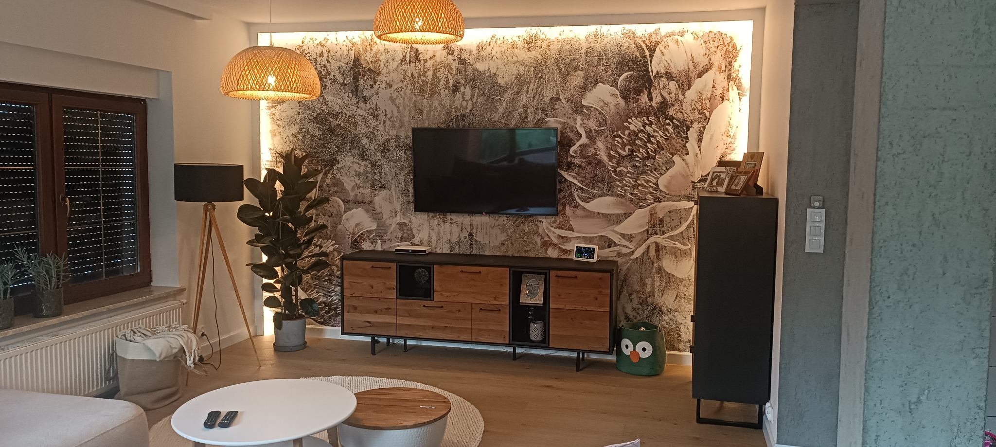 Nowoczesny salon z tapetą w motywy kwiatowe, drewnianą komodą pod telewizorem, lampami z abażurami z wikliny i zielonym koszem w kształcie sowy.