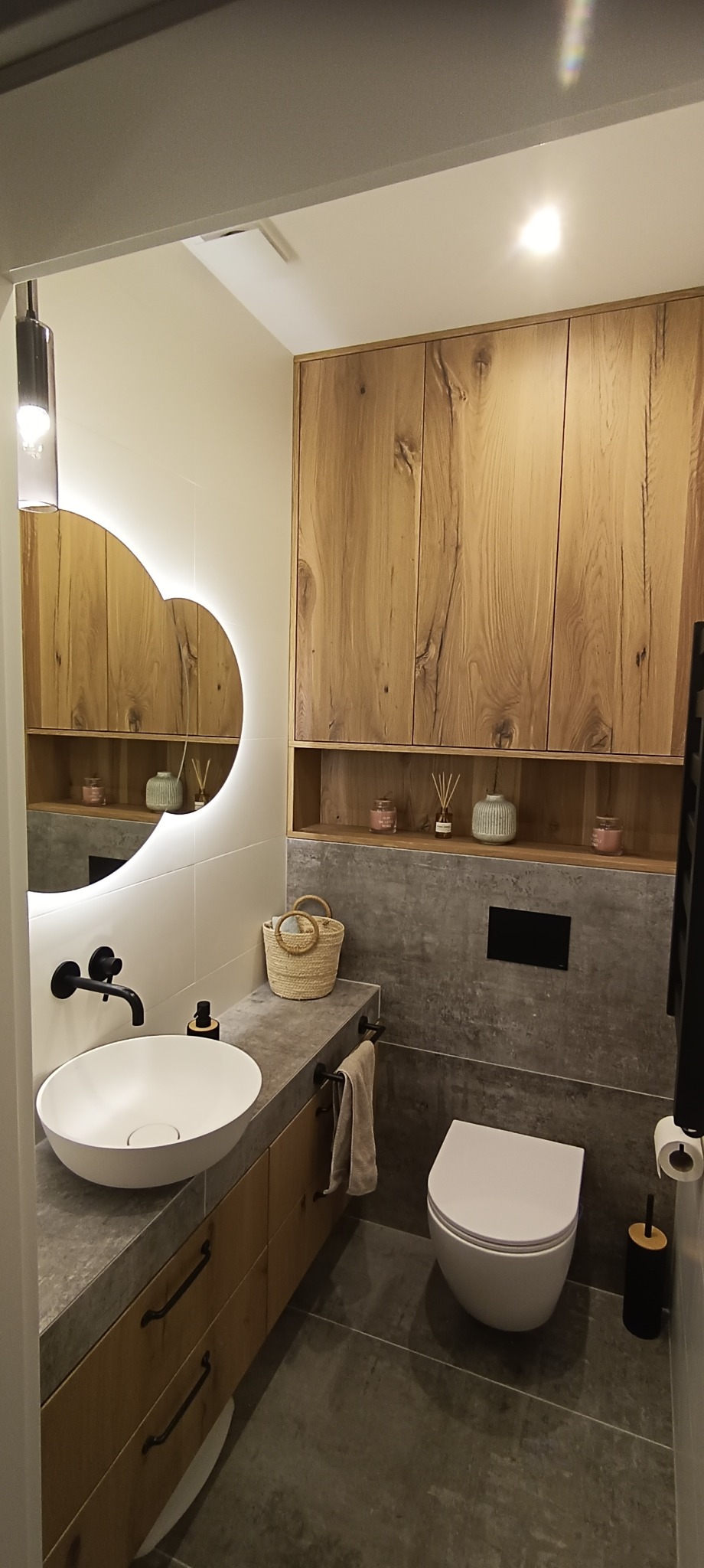 Nowoczesna łazienka z okrągłym lustrem z podświetleniem LED, drewnianymi szafkami, umywalką nablatową i szarą płytką imitującą beton.