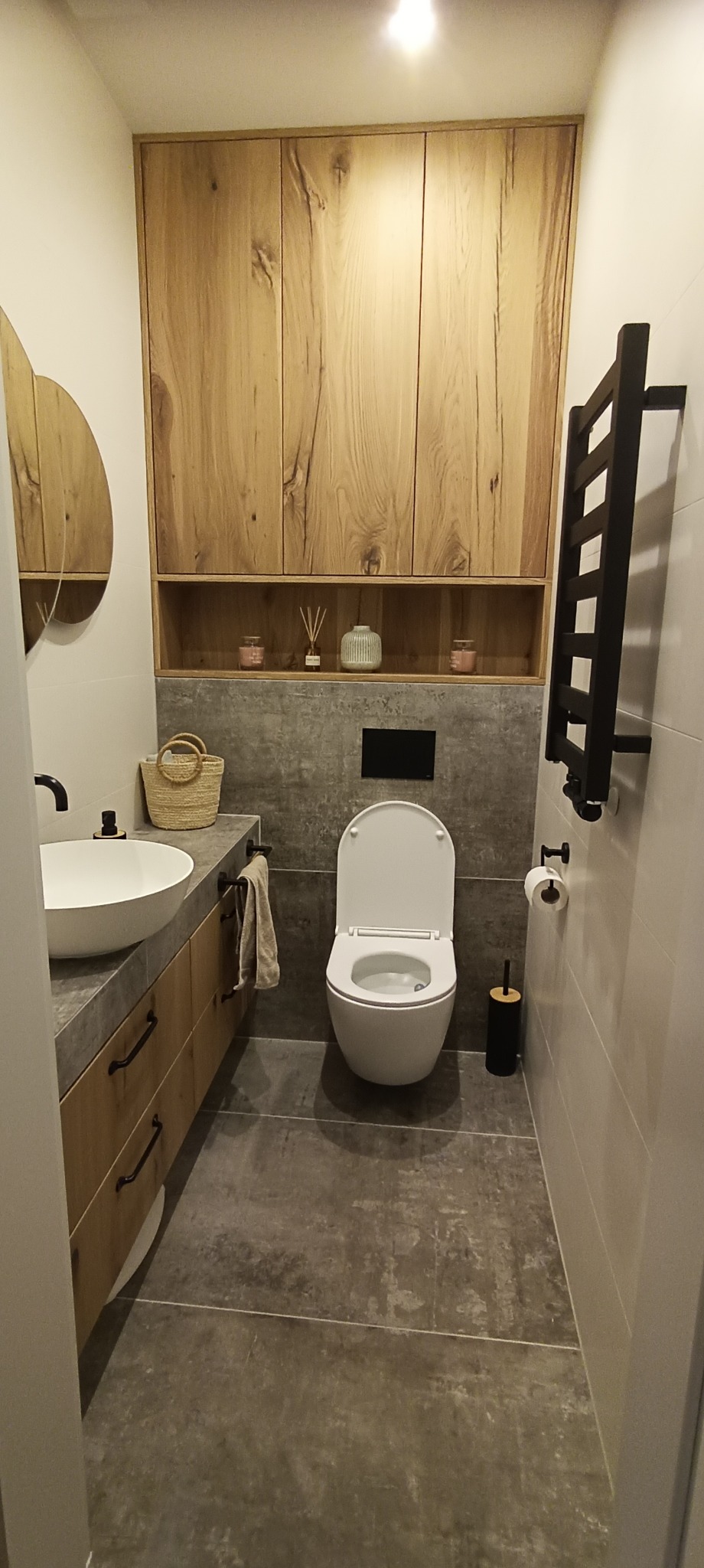 Nowoczesna łazienka z wiszącą toaletą, umywalką nablatową na szafce z drewnianymi frontami i czarnym grzejnikiem drabinkowym, wykończona w szarościach i drewnie.