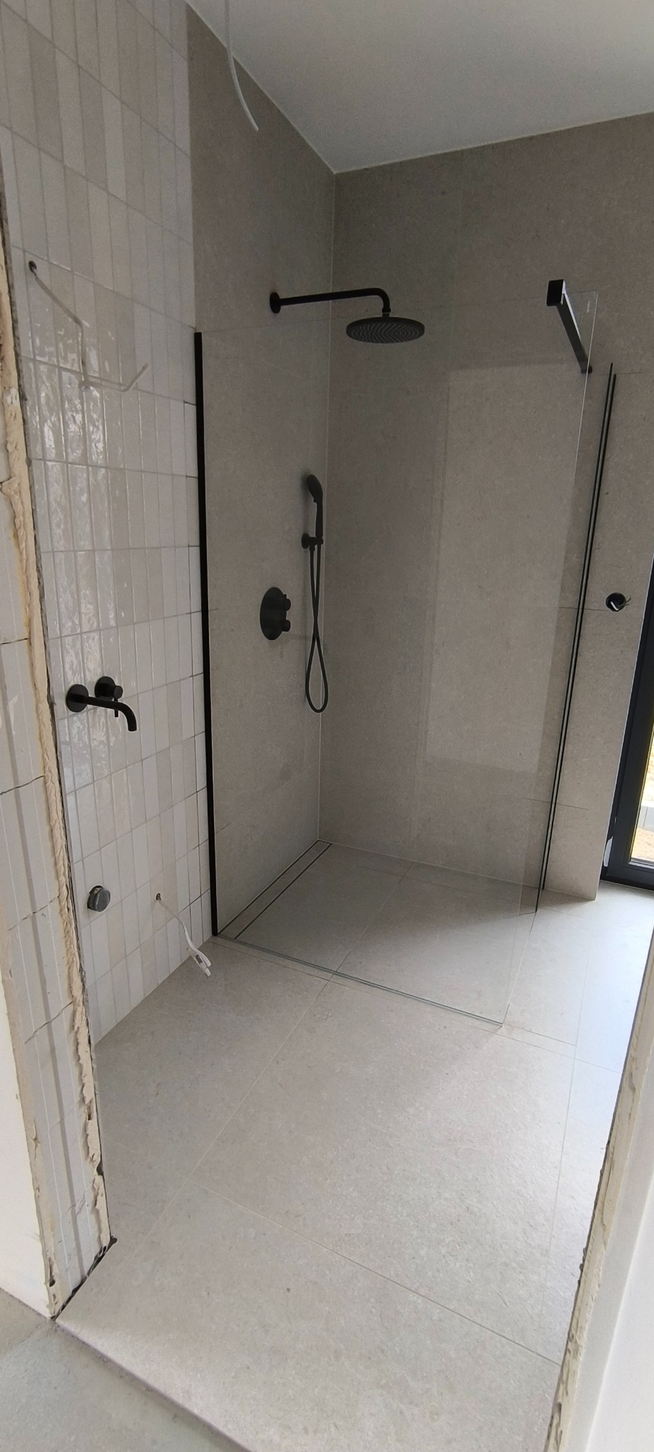 Nowoczesna kabina prysznicowa typu walk-in z czarną armaturą w trakcie montażu, widoczne niedokończone obrzeża płytek przy wejściu.