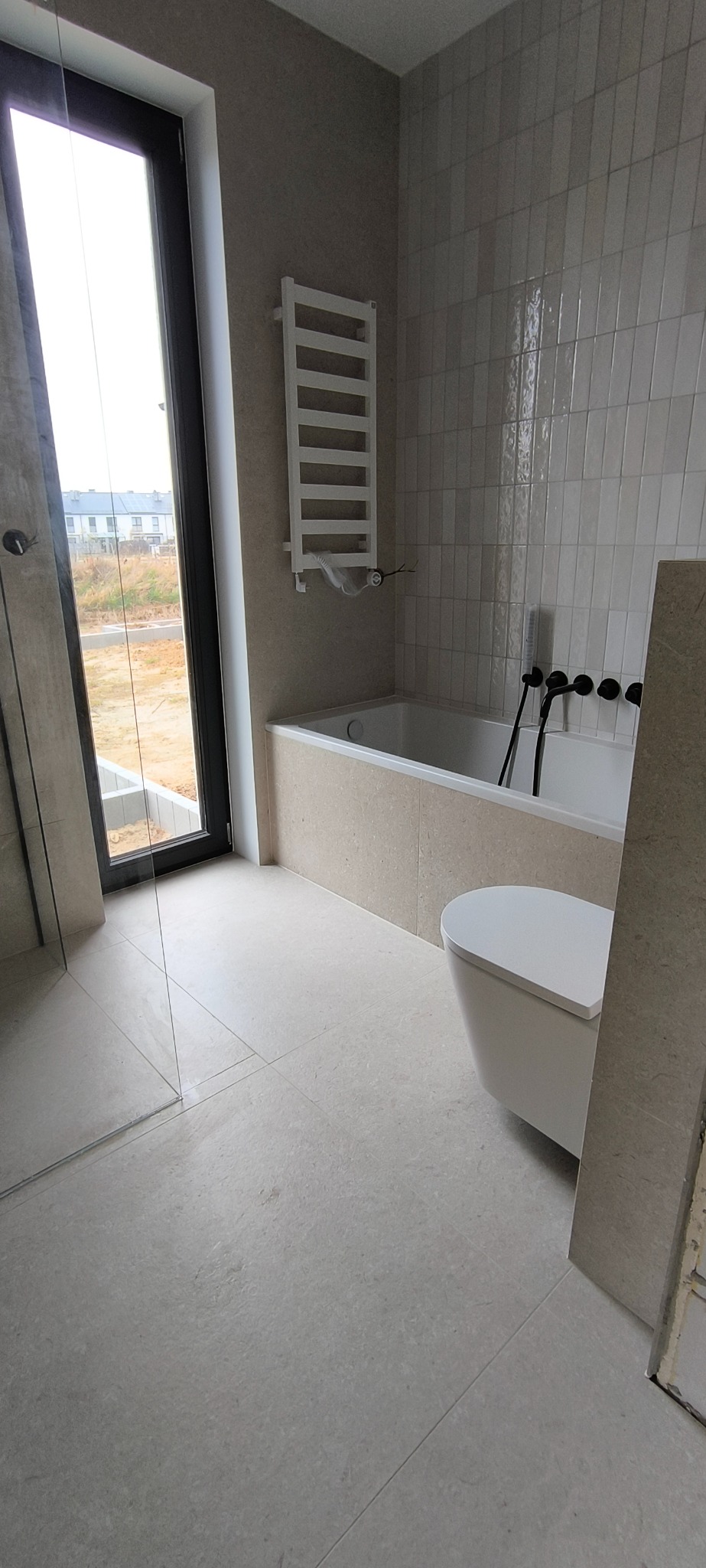 Nowoczesna łazienka w odcieniach beżu z wanną obudowaną płytkami, podwieszaną toaletą, grzejnikiem drabinkowym, czarną armaturą i szklaną kabiną prysznicową, widok na zewnątrz przez duże okno.