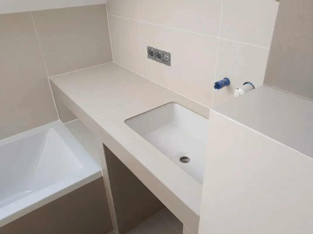 Nowoczesna łazienka w beżowych płytkach z zabudowaną wanną i umywalką, widoczne przyłącza wodne i gniazdka elektryczne przed montażem armatury.