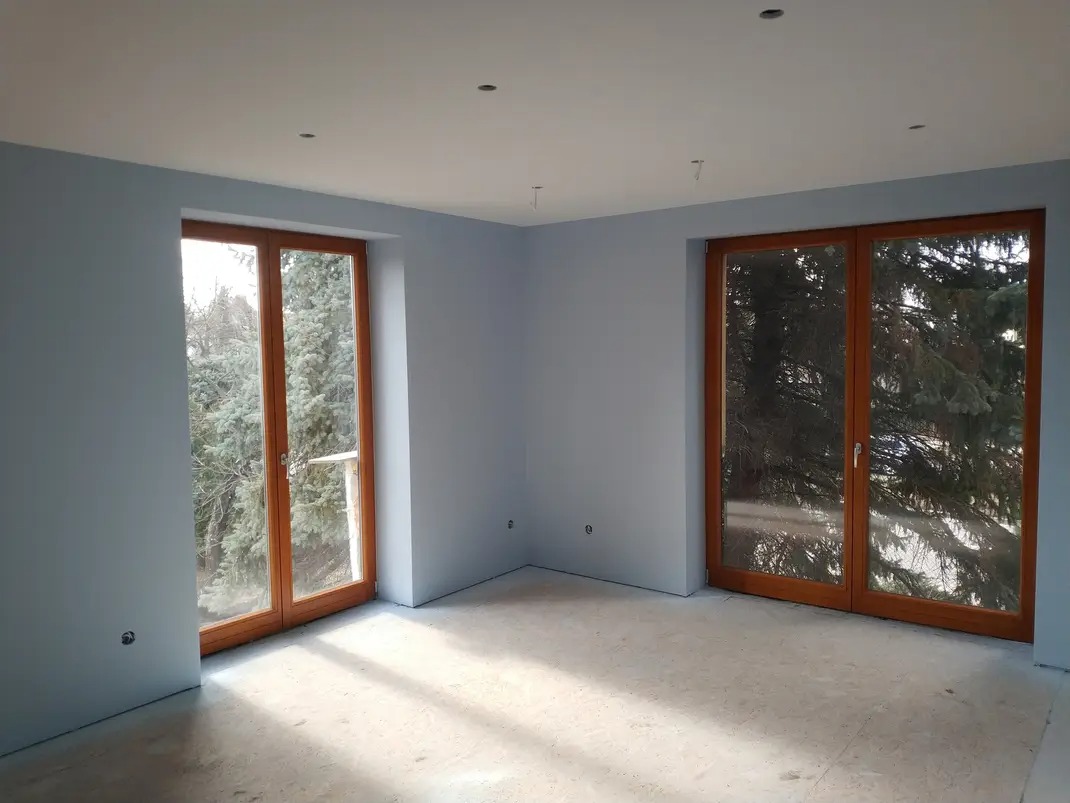 Pomalowane na jasnoniebiesko wnętrze z dwoma drewnianymi oknami balkonowymi, surowa podłoga, widoczne niedokończone prace.