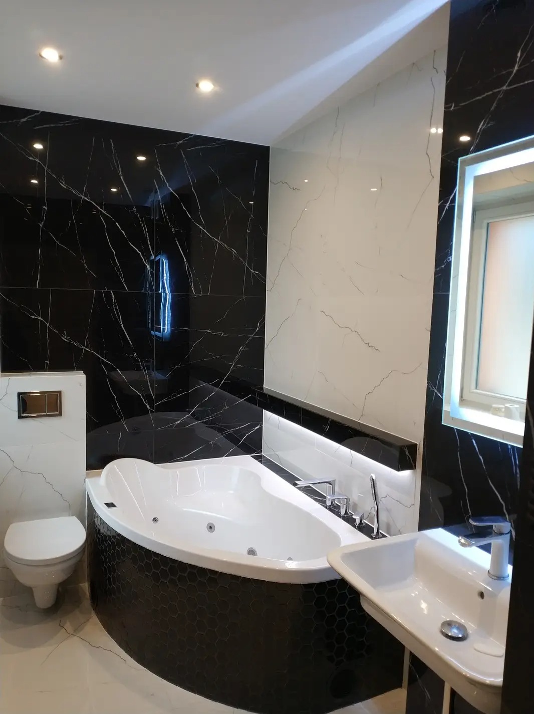 Nowoczesna łazienka z czarno-białymi płytkami imitującymi marmur, narożną wanną z hydromasażem obudowaną czarną mozaiką heksagonalną i podświetlanym lustrem.