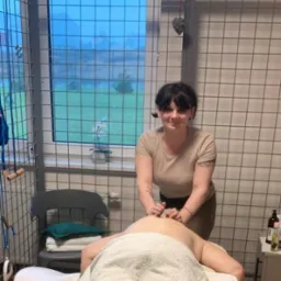 Kobieta wykonuje masaż pleców klientowi na łóżku w gabinecie z kratami w oknach. Na stole butelki z olejkami. Widoczny fragment okna z widokiem na zewnątrz.