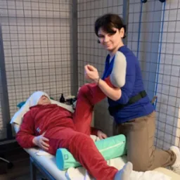 Osoba w czerwonym dresie leży na leżance, a kobieta w niebieskiej bluzce wykonuje ćwiczenia nogi, w tle metalowa klatka i akcesoria do ćwiczeń.