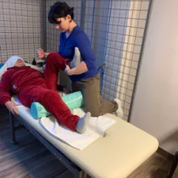 Kobieta w niebieskiej bluzce wykonuje mobilizację stawu kolanowego pacjenta leżącego na leżance rehabilitacyjnej z zielonym wałkiem pod kolanami.