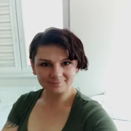 Portret uśmiechniętej kobiety o krótkich, brązowych włosach w zielonej koszulce na tle jasnego pokoju z oknem i grzejnikiem. Widoczny tatuaż na ramieniu.