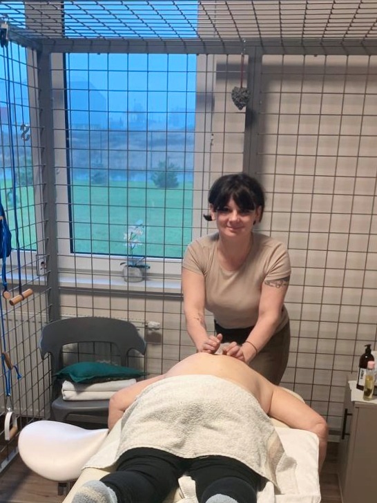 Kobieta wykonuje masaż pleców klientowi na łóżku w gabinecie z kratami w oknach. Na stole butelki z olejkami. Widoczny fragment okna z widokiem na zewnątrz.