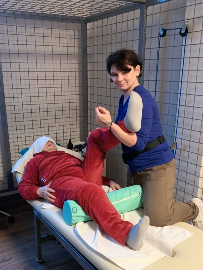 Osoba w czerwonym dresie leży na leżance, a kobieta w niebieskiej bluzce wykonuje ćwiczenia nogi, w tle metalowa klatka i akcesoria do ćwiczeń.