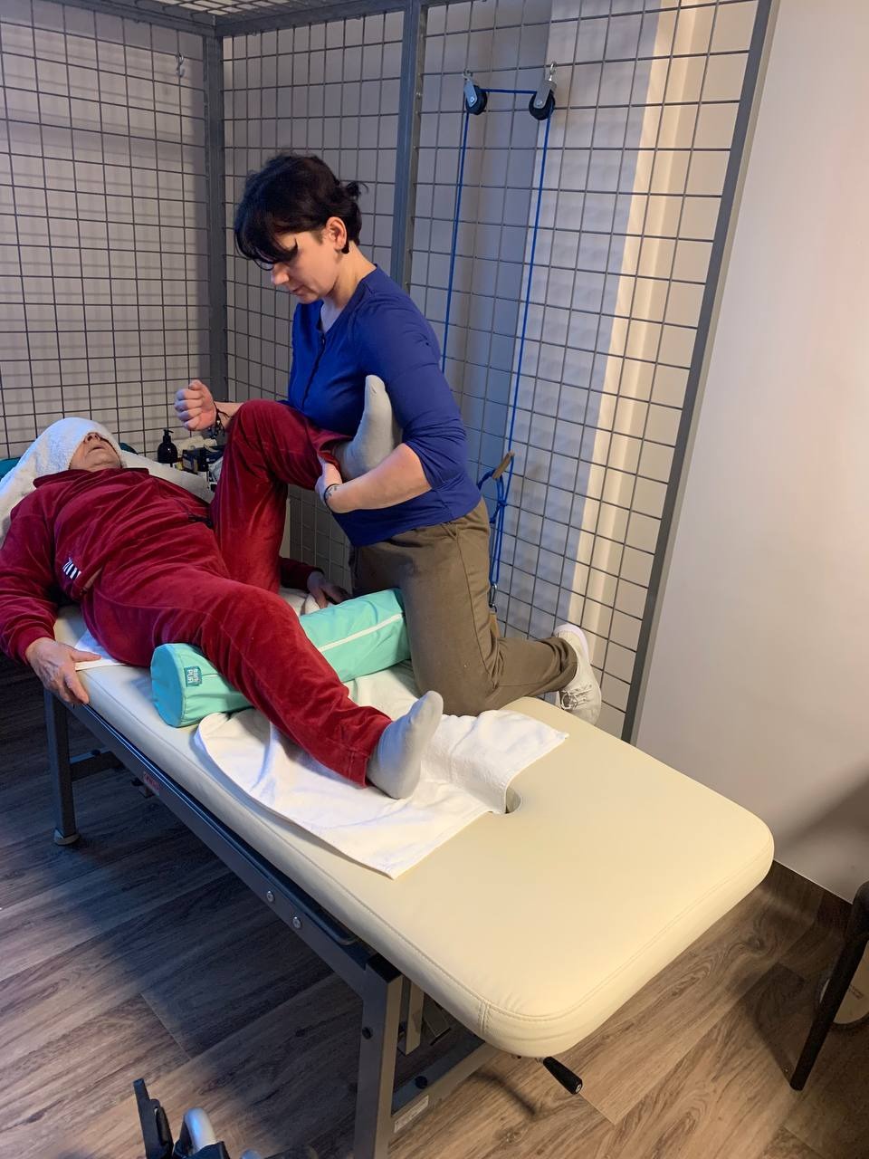 Kobieta w niebieskiej bluzce wykonuje mobilizację stawu kolanowego pacjenta leżącego na leżance rehabilitacyjnej z zielonym wałkiem pod kolanami.