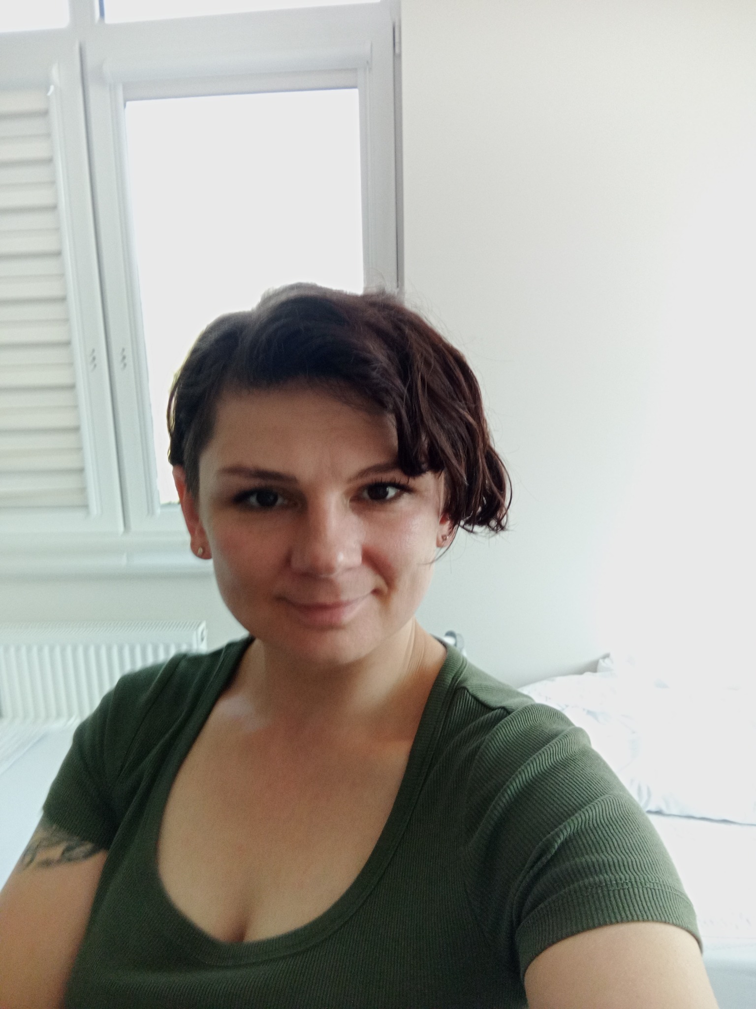 Portret uśmiechniętej kobiety o krótkich, brązowych włosach w zielonej koszulce na tle jasnego pokoju z oknem i grzejnikiem. Widoczny tatuaż na ramieniu.