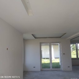 Usługi remontowe Tomasz Stańczyk - Wnętrze pomieszczenia w stanie deweloperskim, ściany przygotowane pod malowanie, widoczne kable elektryczne, sufit z podwieszaną konstrukcją, duże okno balkonowe.