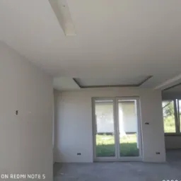 Wnętrze pomieszczenia w stanie deweloperskim, ściany przygotowane pod malowanie, widoczne kable elektryczne, sufit z podwieszaną konstrukcją, duże okno balkonowe.