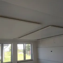 Wykończony sufit podwieszany z płyt kartonowo-gipsowych w jasnym pomieszczeniu z oknami, widoczne przewody oświetleniowe, drabina w tle.