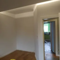 Pomieszczenie po malowaniu z drewnianą podłogą, widoczny fragment żółtego odkurzacza i kabel na podłodze, w tle otwarte drzwi z zabezpieczoną folią podłogą.