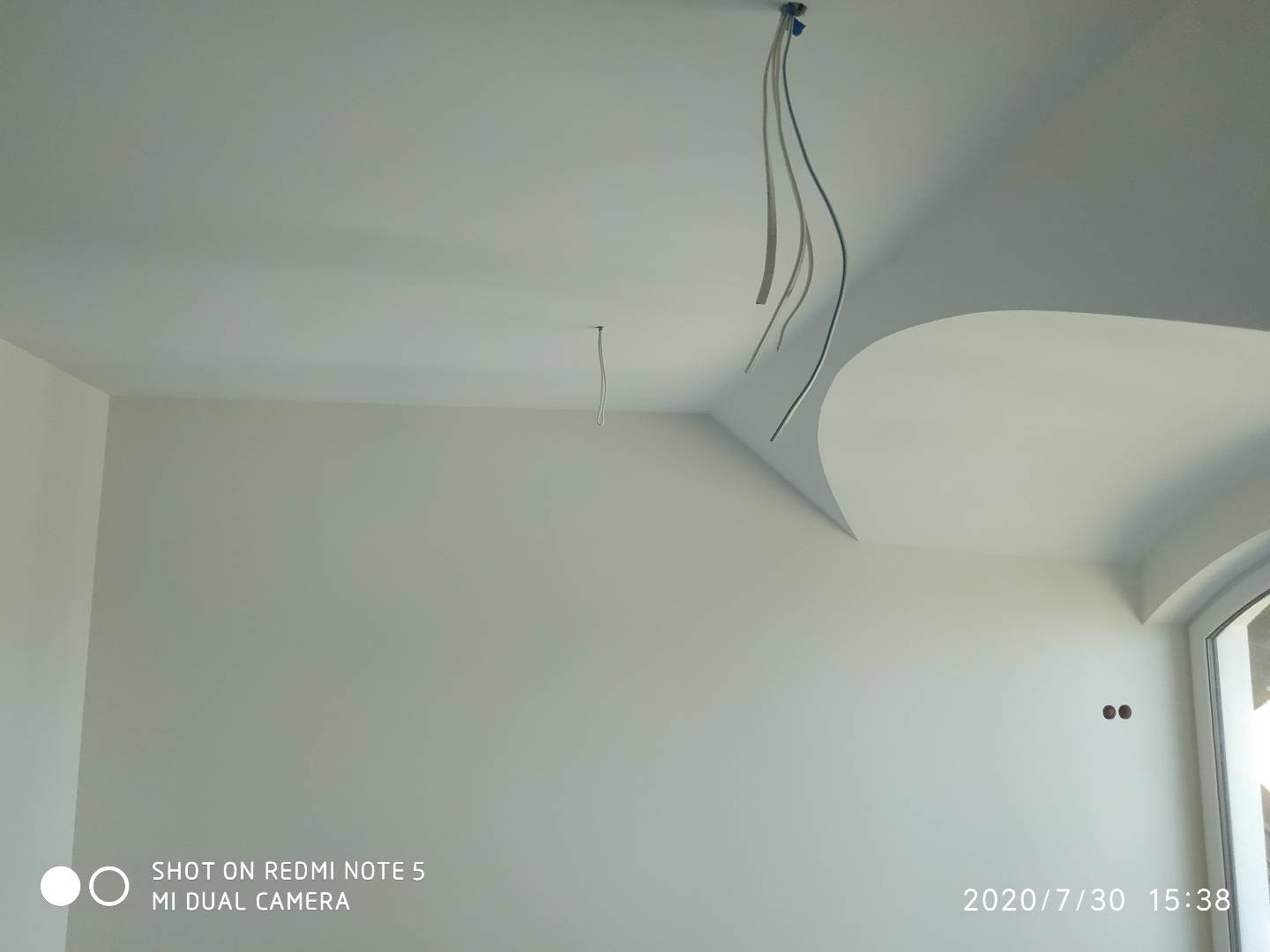 Białe, świeżo pomalowane wnętrze w Skierniewicach z dekoracyjnym elementem sufitowym i wystającymi przewodami elektrycznymi, widoczne otwory na gniazdka elektryczne na ścianie.