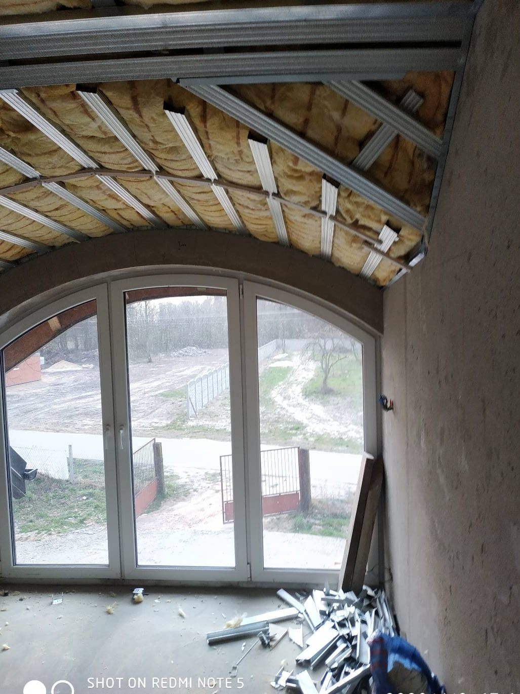 Sufit podwieszany w trakcie montażu z widoczną izolacją i metalową konstrukcją, duże okno z widokiem na zewnątrz, resztki materiałów na podłodze.