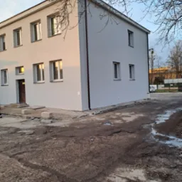 Dwukondygnacyjny budynek po świeżym malowaniu elewacji na szaro, widoczne nieliczne elementy wykończeniowe wokół budynku i błotniste podłoże.