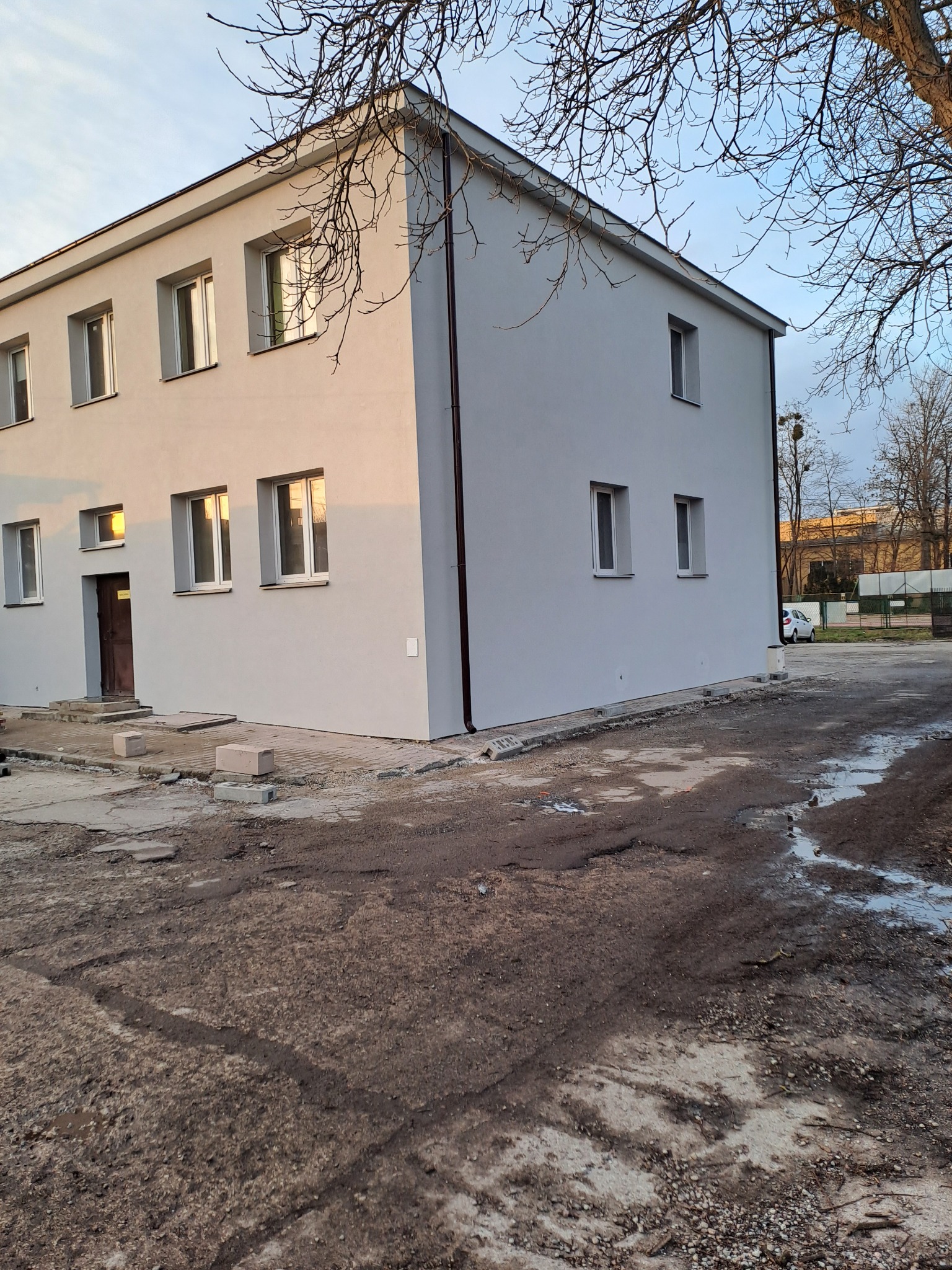 Dwukondygnacyjny budynek po świeżym malowaniu elewacji na szaro, widoczne nieliczne elementy wykończeniowe wokół budynku i błotniste podłoże.