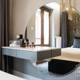 Toaletka z szufladami w szaro-białej kolorystyce, odbicie w okrągłym lustrze ukazuje fragment sypialni z ł&oacute;żkiem i lampami wiszącymi, widoczne flakony perfum i szkatułka na blacie, logo firmy...