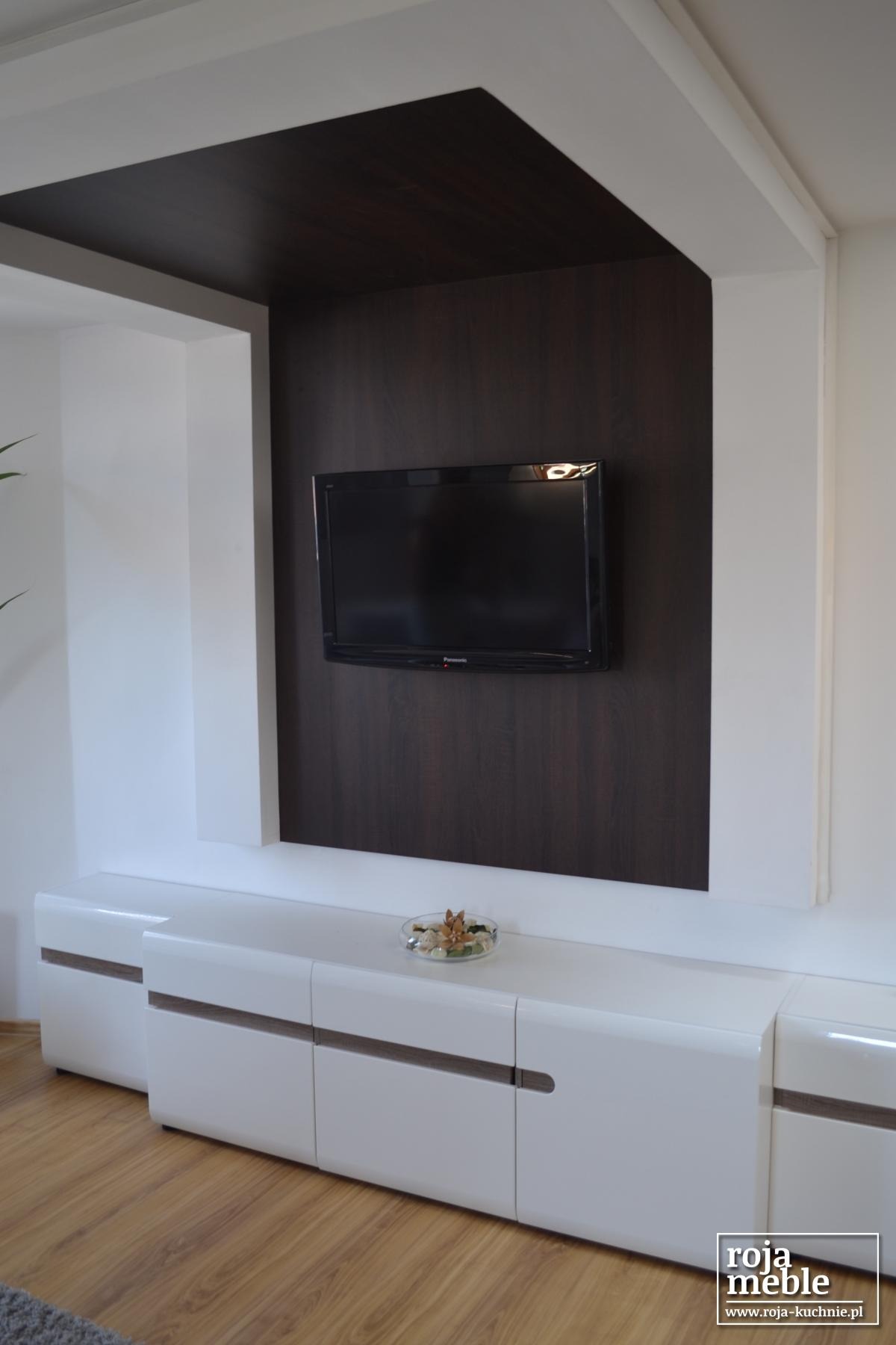 Nowoczesna szafka RTV z białymi, lakierowanymi frontami i ciemnobrązowym panelem ściennym pod telewizor, dekoracja w szklanej miseczce na blacie.
