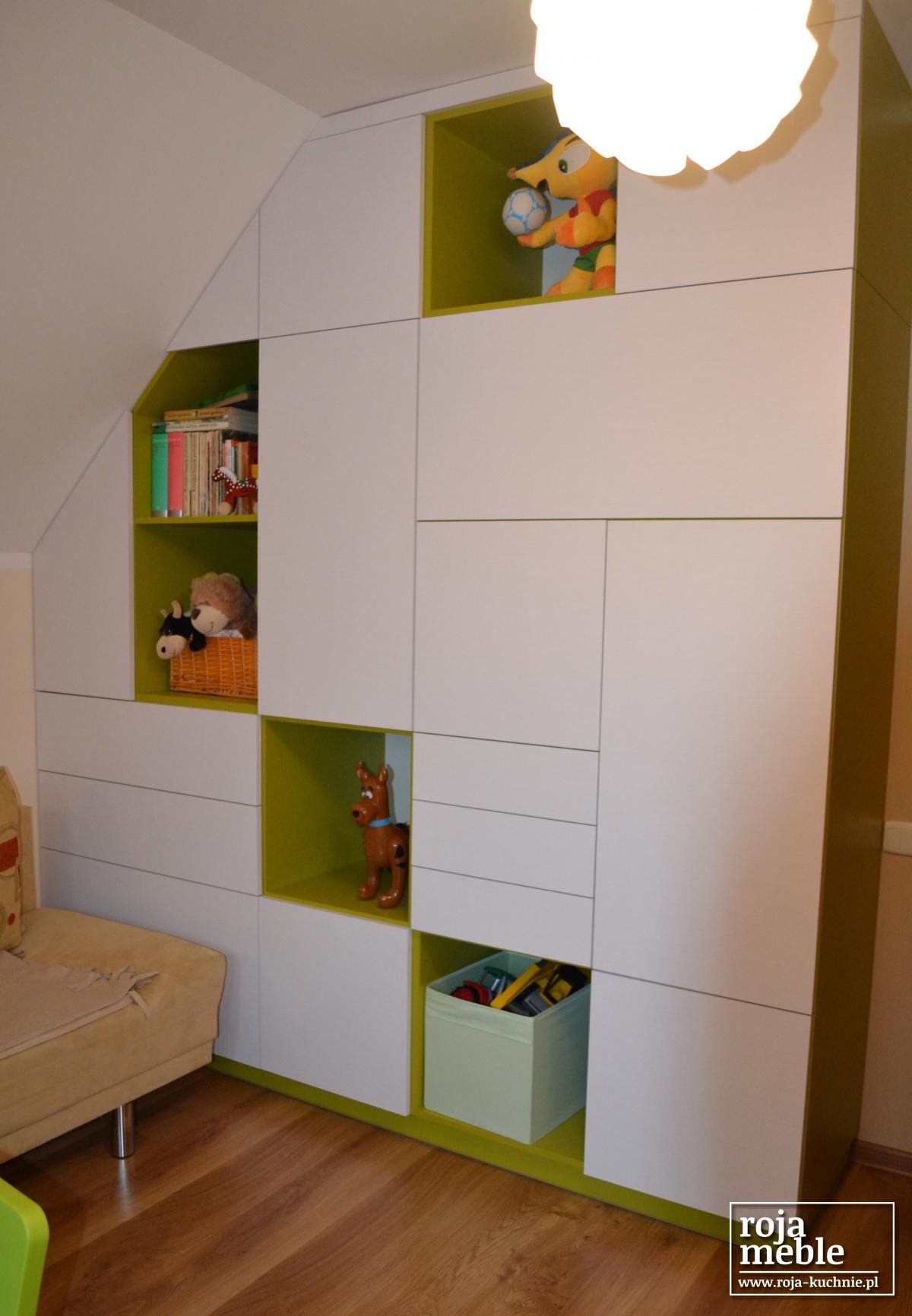 Biała, modułowa zabudowa meblowa w pokoju dziecięcym, z zielonymi wnękami wypełnionymi zabawkami i książkami, dopasowana do skosu dachu, oświetlona lampą sufitową.
