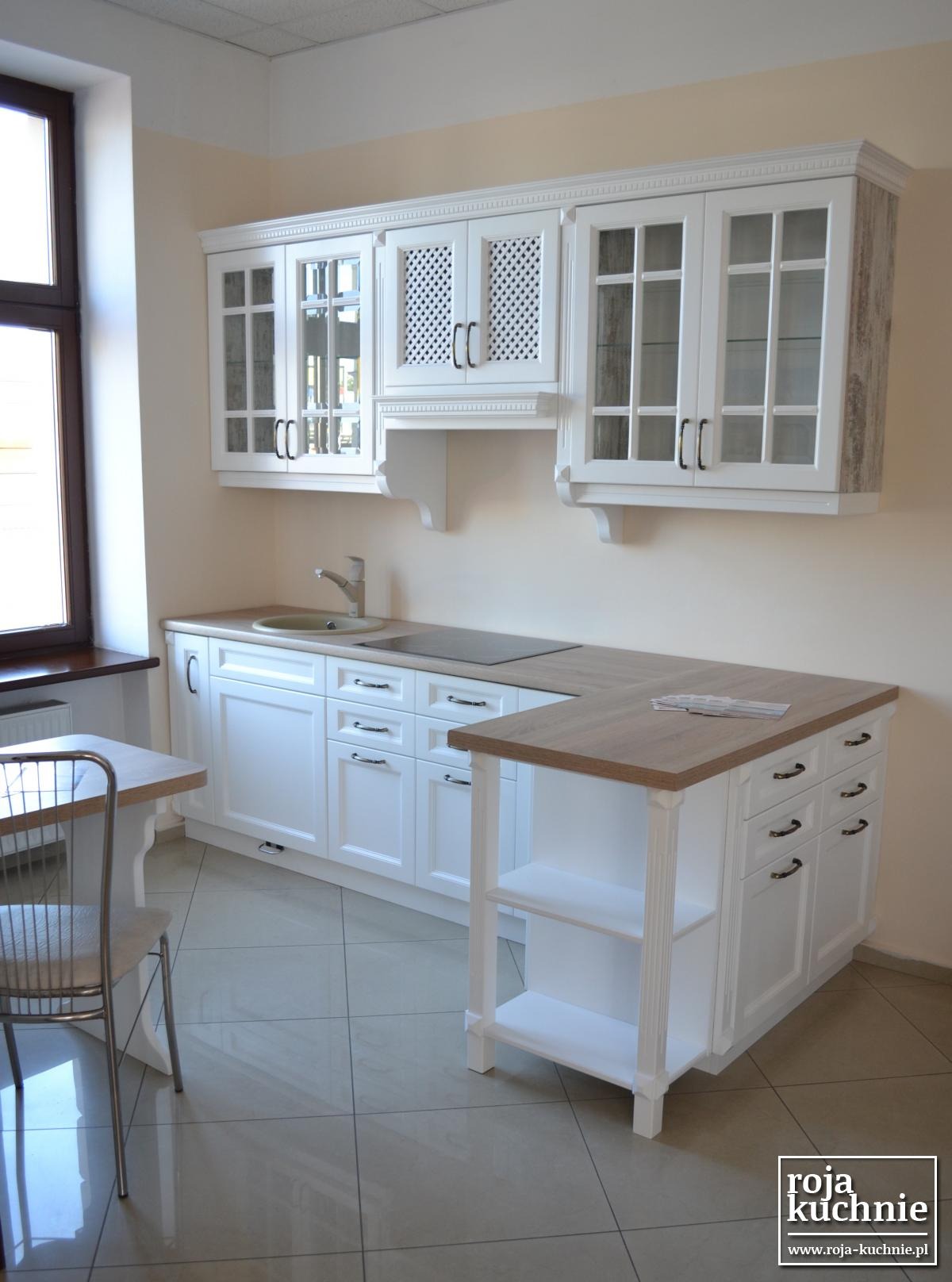 Jasna kuchnia z białymi szafkami z przeszklonymi frontami i drewnianymi blatami, widok na okno i stół z krzesłem, logo firmy w rogu.