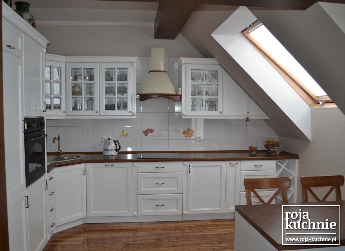 Jasna kuchnia w stylu rustykalnym z białymi szafkami, drewnianym blatem i oknem dachowym, widoczne szklane witryny z dekoracyjnymi naczyniami i drewniany stół z krzesłami.