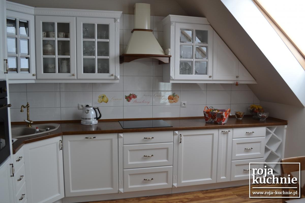 Biała kuchnia z drewnianym blatem w poddaszu, przeszklone szafki wiszące z dekoracyjnymi elementami, płytki ceramiczne z motywem owoców, czajnik elektryczny i koszyk na owoce na blacie.