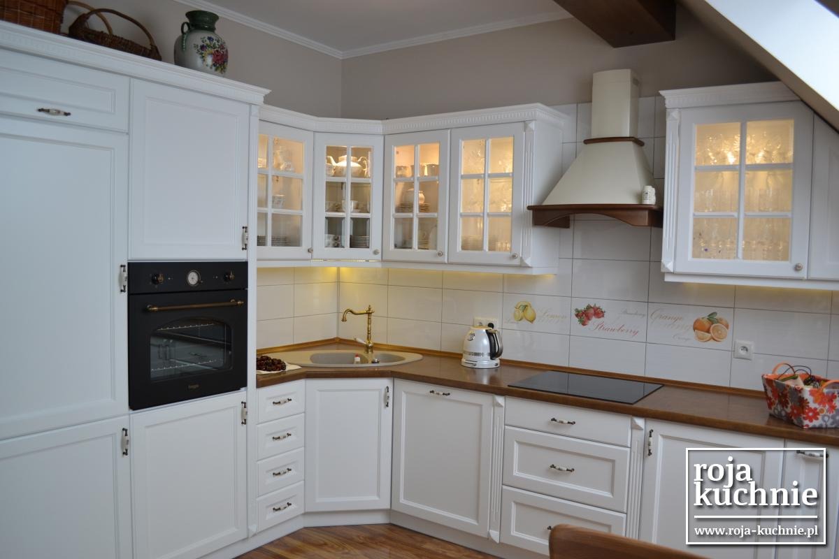 Klasyczna kuchnia z białymi szafkami, drewnianym blatem i czarnym piekarnikiem w zabudowie. Górne szafki przeszklone, podświetlane od wewnątrz. Nad blatem płytki ceramiczne z motywami owoców.