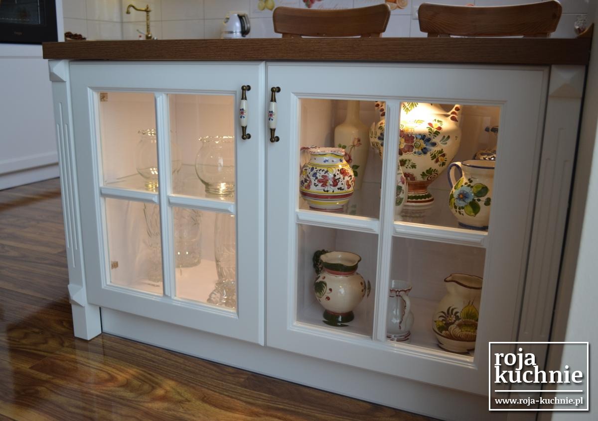 Biała szafka kuchenna z przeszklonymi drzwiczkami, eksponująca kolekcję ozdobnych wazonów i dzbanków w stylu rustykalnym, z drewnianym blatem i ceramicznymi uchwytami z motywem kwiatowym.