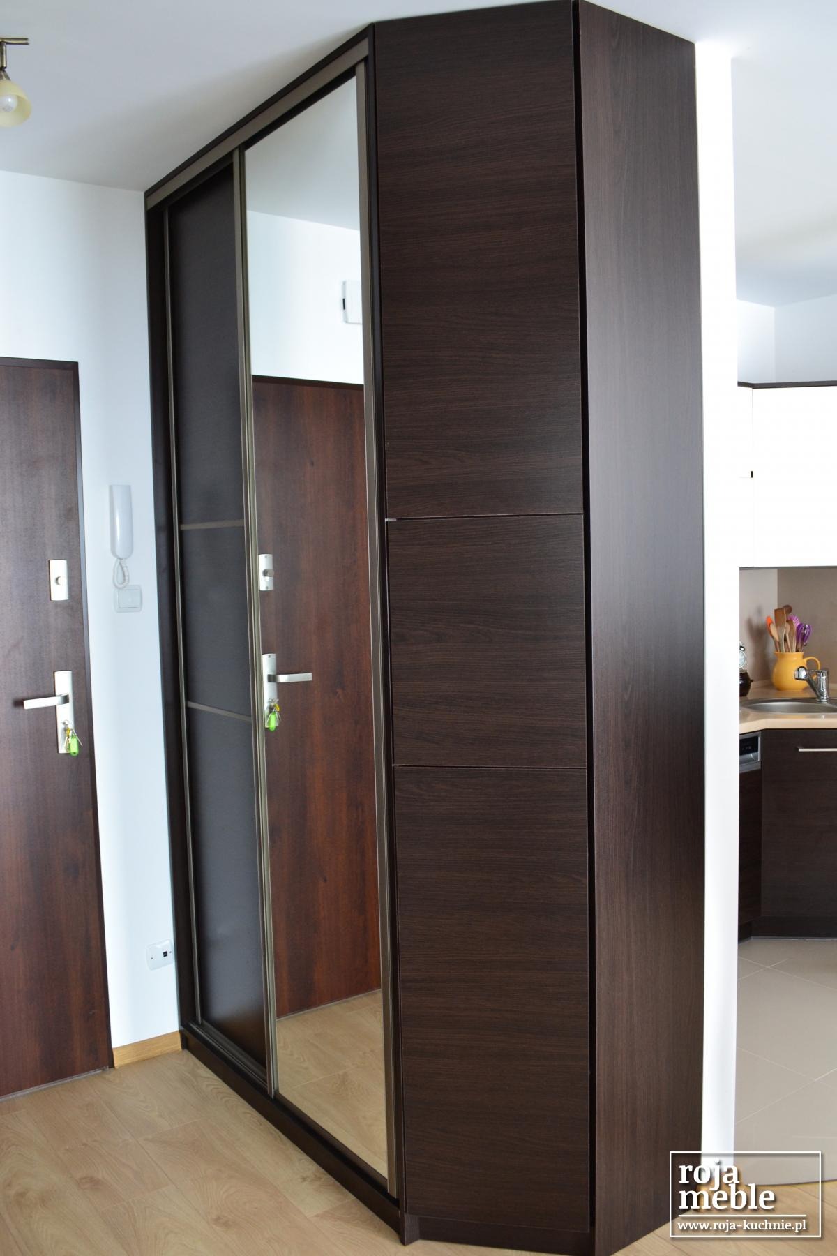 Wysoka szafa wnękowa z przesuwnymi drzwiami z lustrem, w odcieniach ciemnego brązu, widoczna z perspektywy wejścia do mieszkania, z fragmentem kuchni w tle.