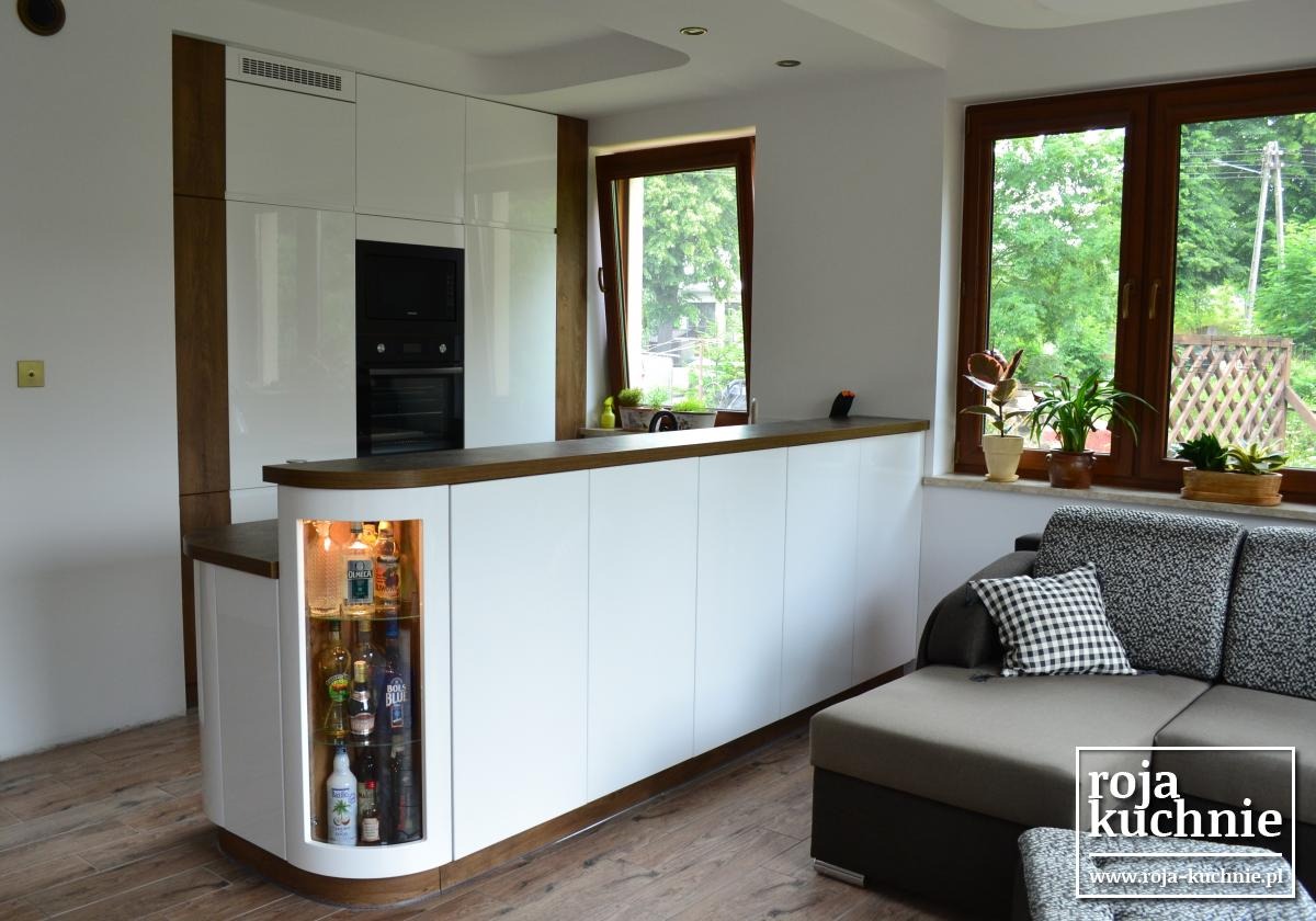 Nowoczesna kuchnia z białymi frontami, drewnianym blatem i podświetlaną półką barową z alkoholem, widoczna z perspektywy salonu z szarą sofą i oknami z widokiem na zieleń.