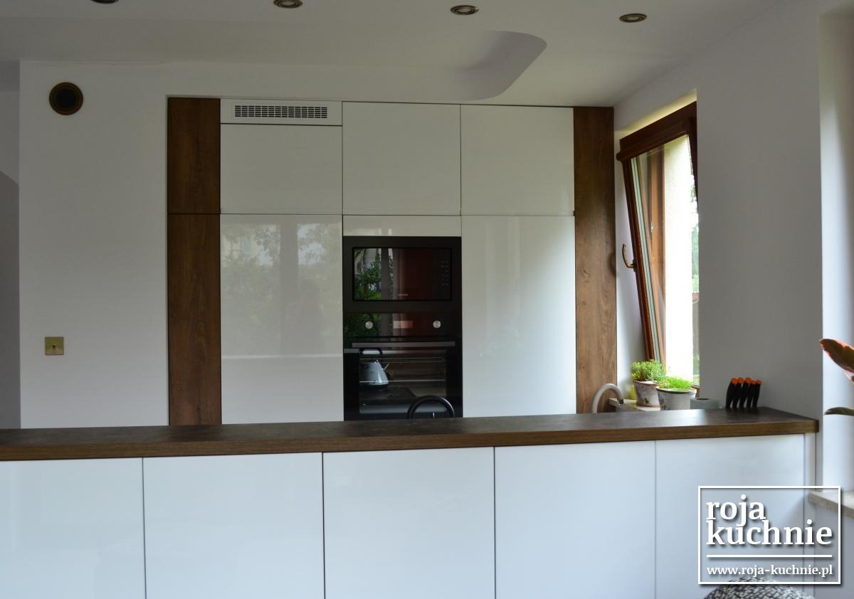 Białe, lakierowane szafki kuchenne z drewnianymi akcentami po bokach, ciemny blat i otwarte okno z ziołami w doniczkach.