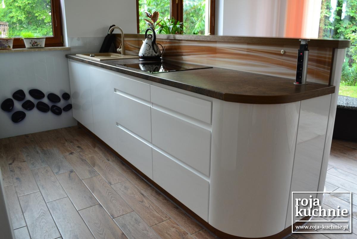 Biała wyspa kuchenna z zaokrąglonymi bokami, brązowym blatem i wbudowaną płytą grzewczą, widok na okno z roślinami, dekoracja ścienna z czarnych kamieni, drewniana podłoga, wysuwane gniazdko...