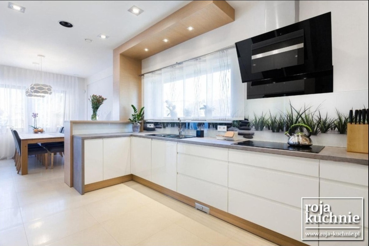 Nowoczesna, jasna kuchnia z białymi, lakierowanymi frontami szafek, szarym blatem, czarnym okapem i drewnianymi akcentami, widok na jadalnię z oknem.