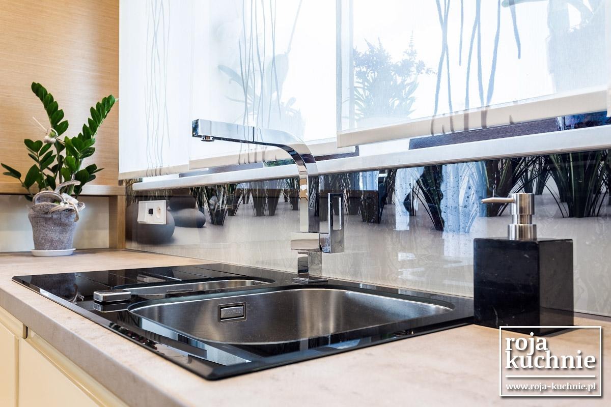 Nowoczesny zlewozmywak wkomponowany w blat kuchenny, bateria z chromowanym wykończeniem, dozownik mydła z czarnego kamienia, roślina doniczkowa i rolety okienne z motywem roślinnym w tle.