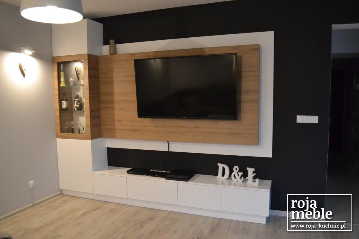 Nowoczesna zabudowa meblowa w salonie z telewizorem, szafką RTV i witryną z oświetleniem, na ścianie dekoracyjne litery D&Ł, logo firmy w prawym dolnym rogu.