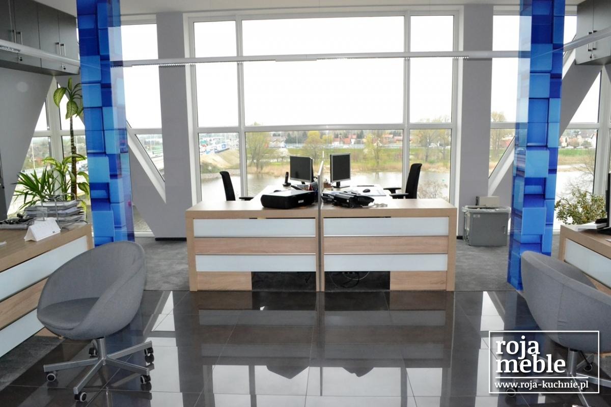 Nowoczesne biuro z dwoma biurkami z jasnego drewna i białymi frontami, krzesła biurowe, duże okna z widokiem na rzekę, szare krzesło obrotowe i dekoracyjny panel z niebieskich kostek.