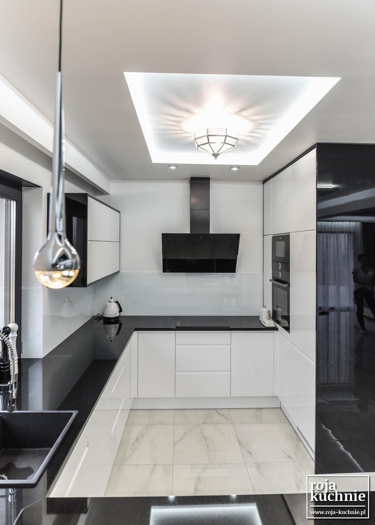 Nowoczesna kuchnia w bieli i czerni z błyszczącymi powierzchniami, oświetleniem sufitowym i marmurową podłogą, widziana z perspektywy szerokokątnej.