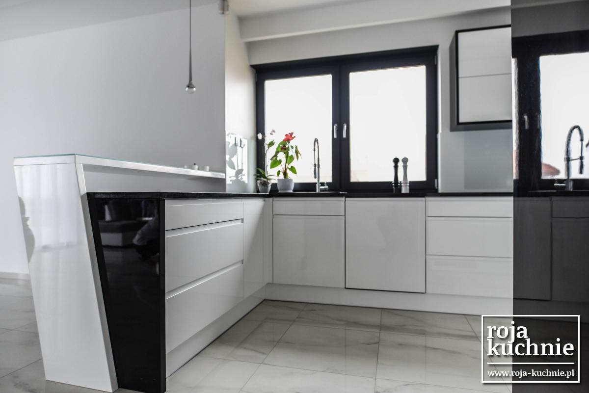 Nowoczesna, jasna kuchnia z białymi frontami szafek, czarnym blatem i dużym oknem z czarną ramą, na blacie stoi doniczka z kwiatem, widoczny fragment wyspy kuchennej z czarnym panelem bocznym.